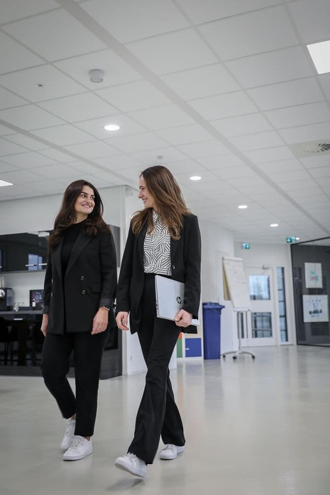 Twee werknemers lopen lachend zij aan zij door de gang, 1 van hen draagt een laptop onder haar arm.