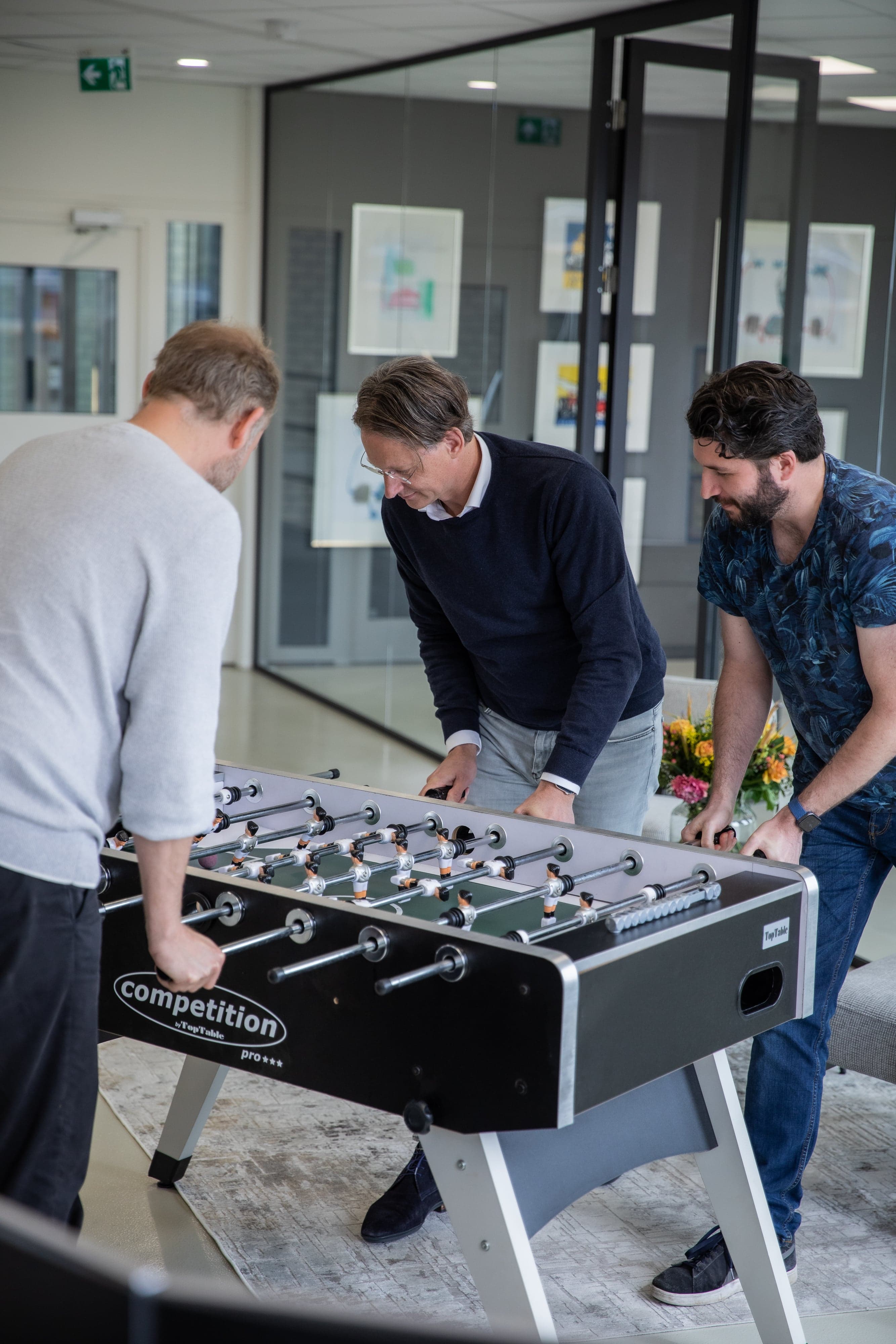 Drie werknemers spelen geconcentreerd tafelvoetbal.