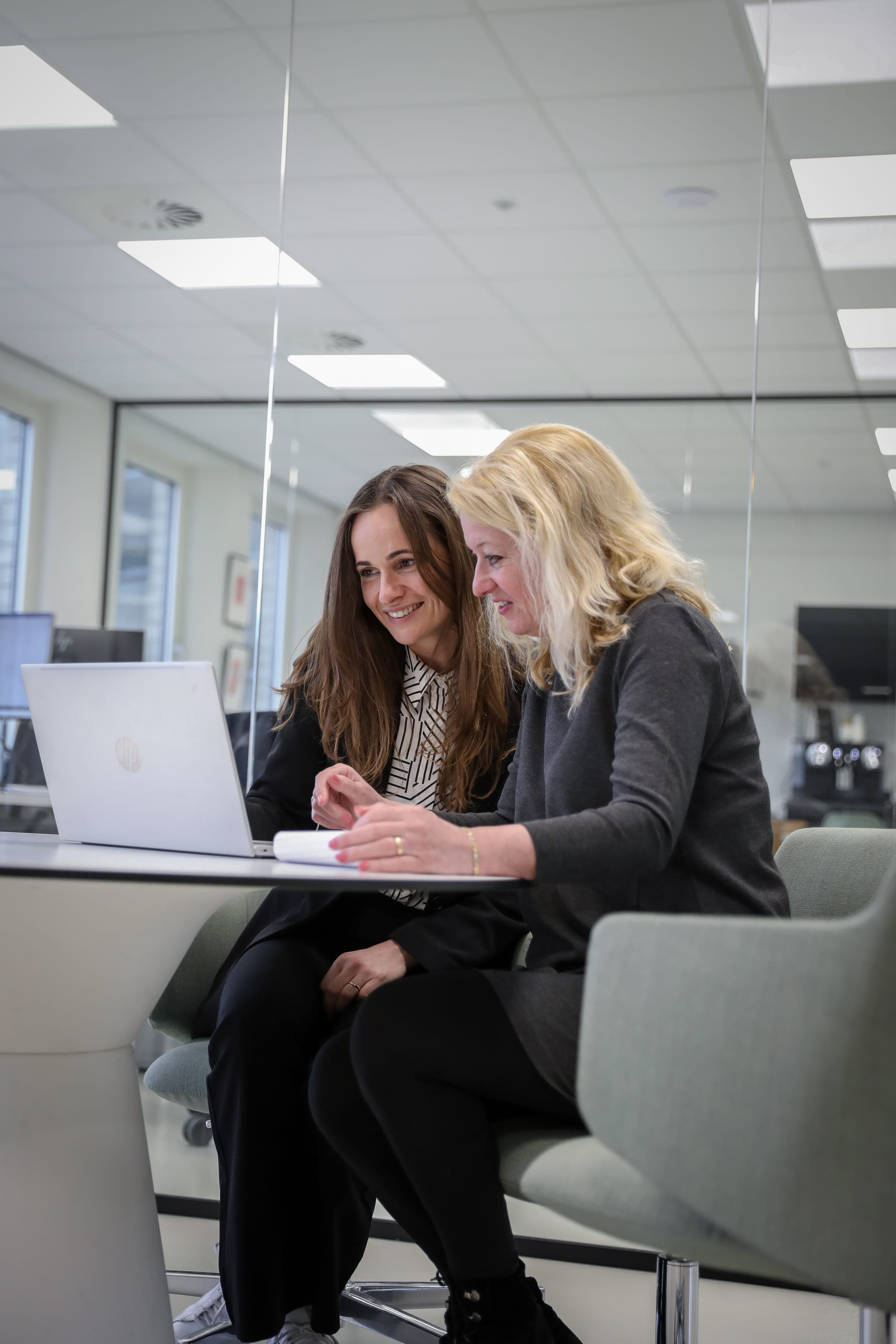 Twee werknemers zitten aan een ronde tafel en glimlachen naar hun laptop.