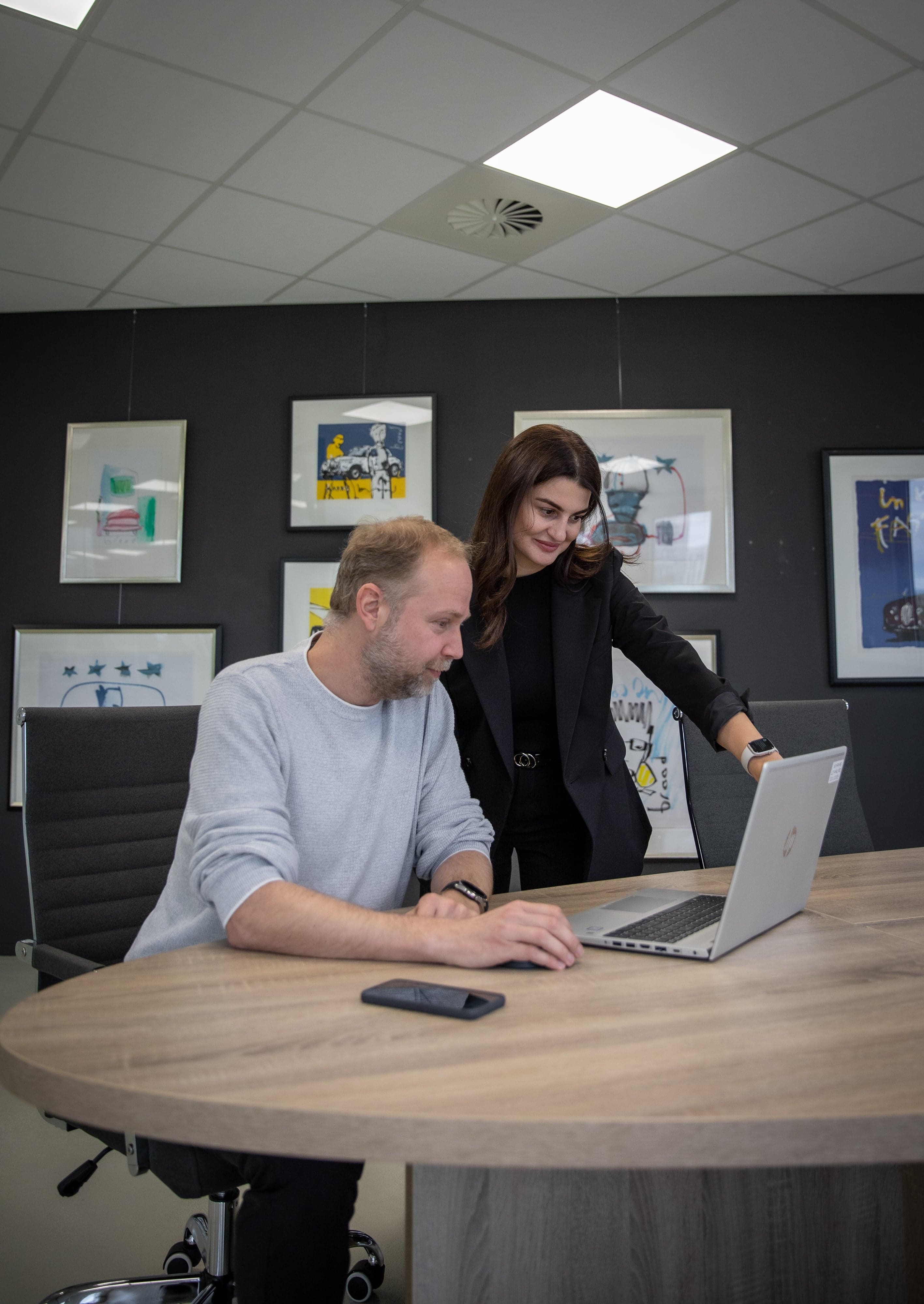 Een klant zit aan tafel met een laptop, terwijl een werknemer staand meekijkt en wijst naar de laptop.