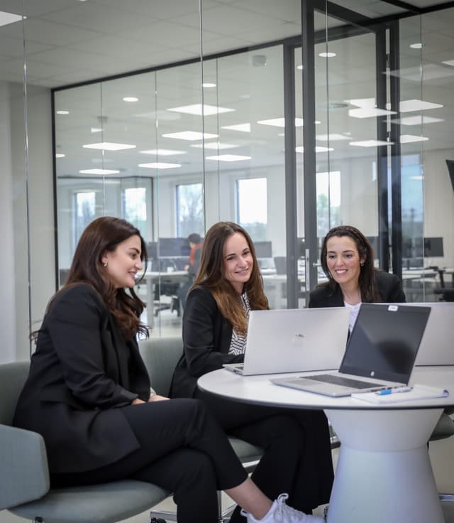 Drie werknemers zitten aan een ronde tafel en kijken met een lach op hun gezicht naar een laptop.