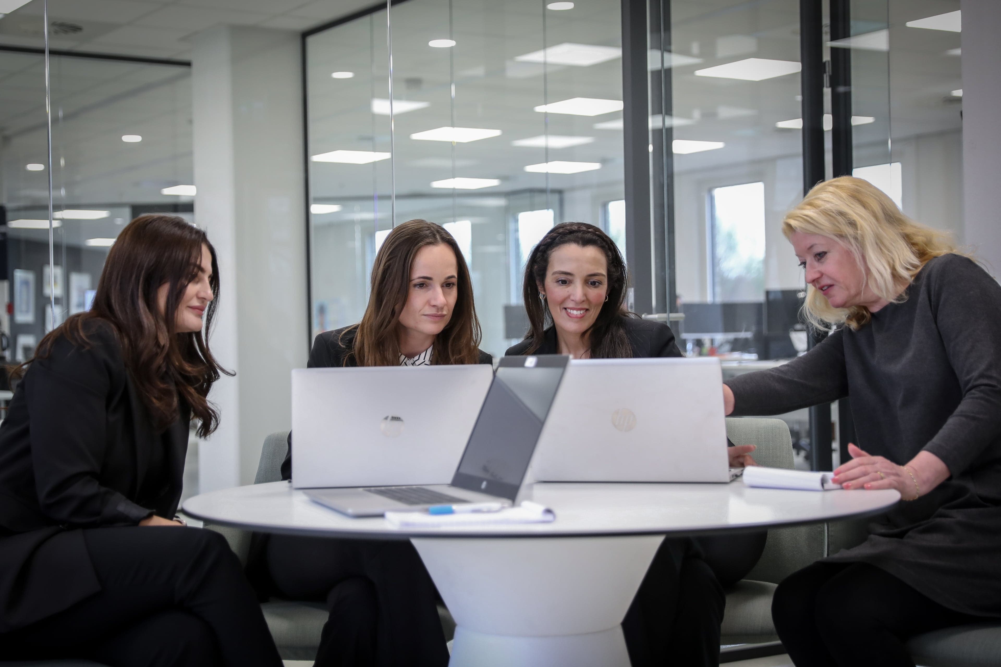 Vier werknemers zitten aan een ronde tafel met laptops terwijl één wijst naar haar scherm.